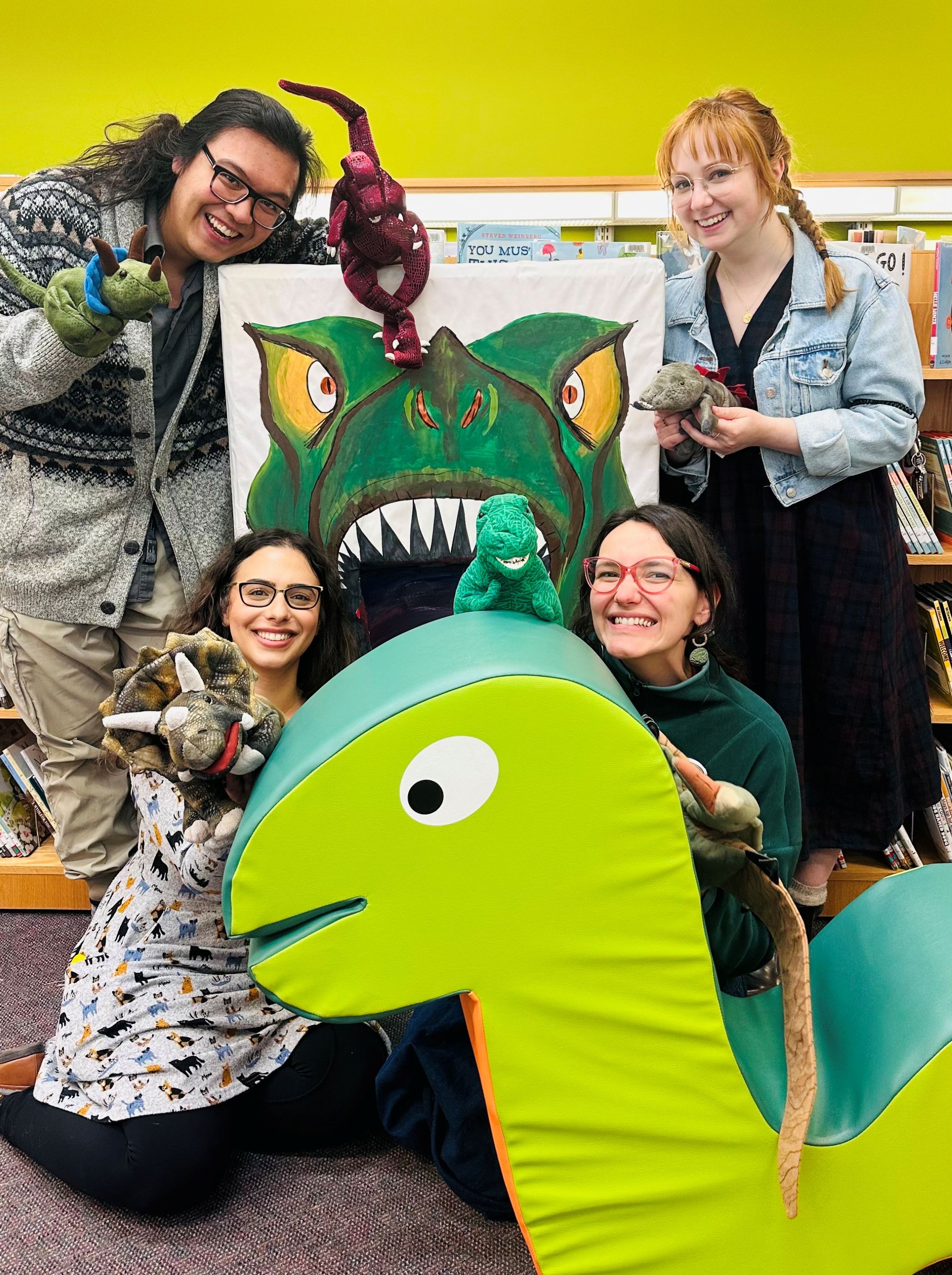 Four people holding hand puppets in front of a large snake-shaped children's chair