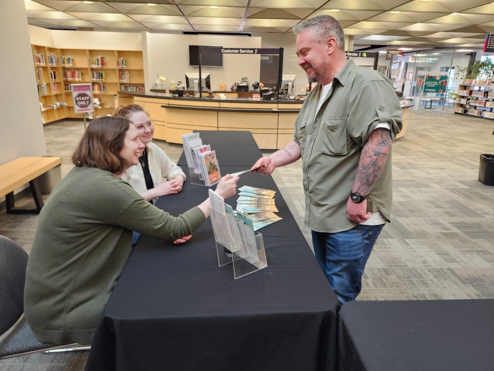Two people sitting at a table and handing a brochure to a person who is standing in front of the table