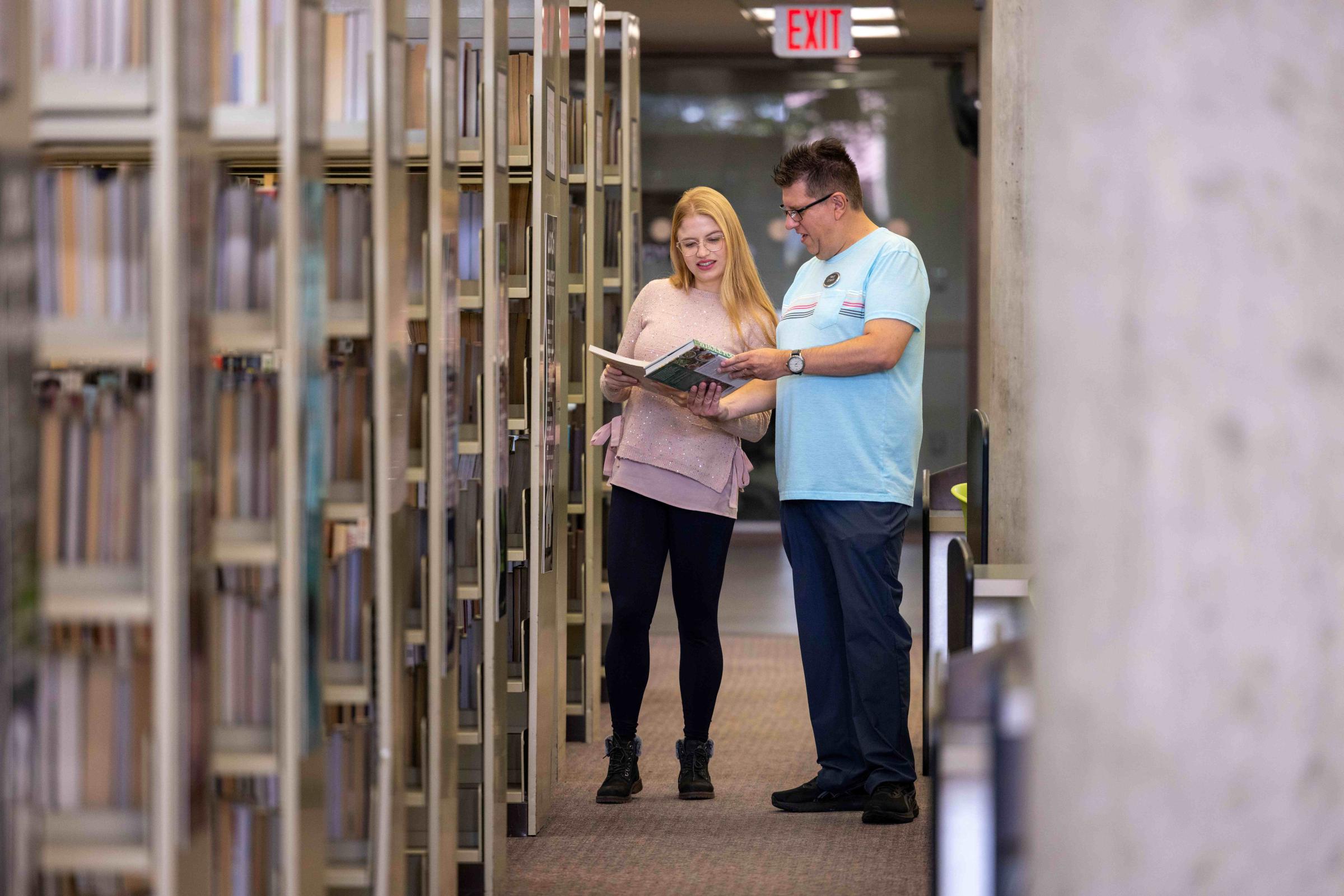 Two adults holding and looking at a book next to a bay of book shelves.  
