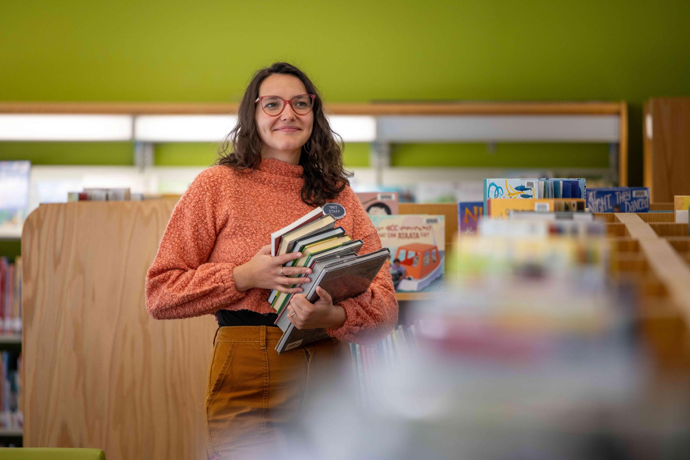 A person holding a stack of books next to a book shelf